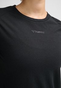 Zbliżenie osoby noszącej czarną sportową koszulkę Hummel z okrągłym dekoltem i małym logo na piersi.