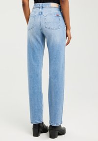 Skats no muguras uz personu, kas valkā gaisvi zilas, augsta jostas taisna silueta džinsas un melnas potīšu zābaciņus ar bloku papēdi uz gaiša fona.