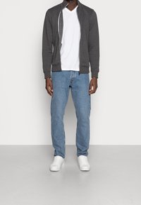 Giacca grigia con zip, maglietta bianca a V, jeans blu e sneakers bianche. La giacca ha tasche laterali e polsini a costine, con una vestibilità casual.