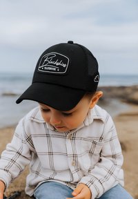 Schwarze Mütze mit einem weißen Logo-Patch, kombiniert mit einem weißen karierten Hemd und blauen Jeans. Das strukturierte Material hebt sich von einem glatten Hintergrund ab.