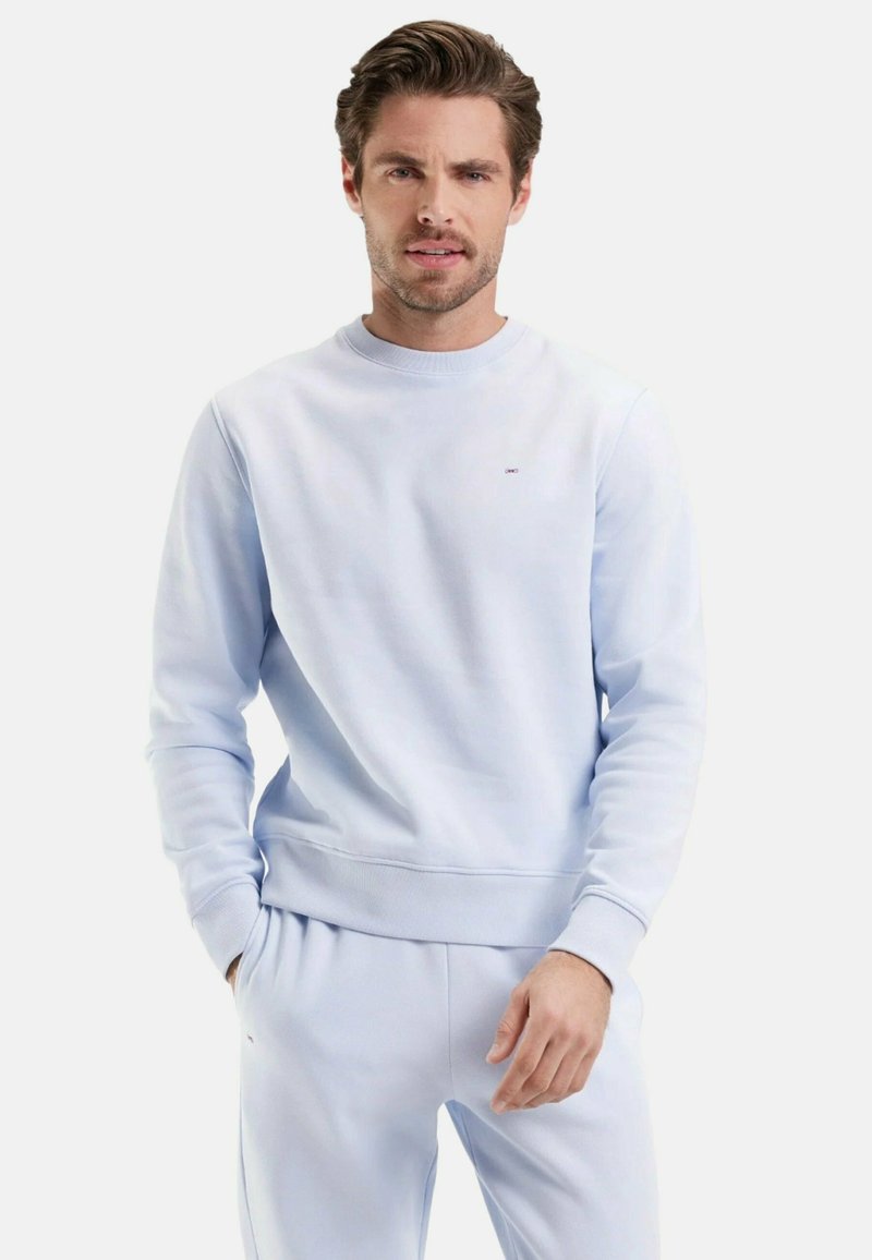 Homme portant un sweat-shirt bleu clair et un pantalon de survêtement assorti, se tenant avec une main dans la poche contre un fond uni.