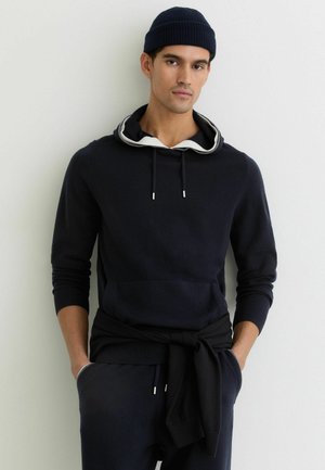 Donkerblauwe hoodie met een gelaagde witte binnenkap, voorzak en zilverkleurige koordaccenten. Gecombineerd met donkerblauwe joggingbroek en een bijpassende beanie.