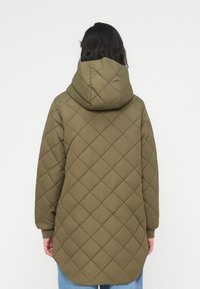 Πράσινο ελιάς καπιτονέ μπουφάν με κουκούλα, με μοτίβο διαμαντιού και ριμπ μανσέτες. Το μήκος φτάνει μέχρι το μέσο του μηρού, με στρογγυλεμένη γραμμή στο τελείωμα.