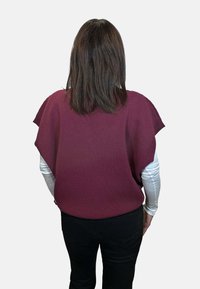 Top in maglia color bordeaux con design ampio simile a un mantello e maniche corte, sovrapposto a una camicia bianca a maniche lunghe, abbinato a pantaloni neri.