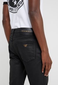 Černé džínové kalhoty s úzkým střihem, zadními kapsami s logem zlatého orla Armani a koženou štítkem na pásku.