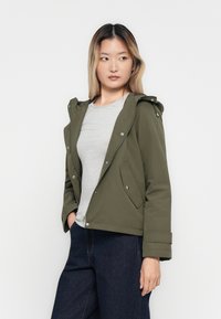 Femme jeune portant une veste à capuche vert olive sur un t-shirt gris et un jean bleu foncé, se tenant les mains dans les poches, regardant sur le côté.