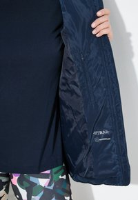 Chaqueta repelente al agua en color marino, con tejido suave y brillante, un bolsillo lateral con cremallera y un logo en el interior cerca del dobladillo.