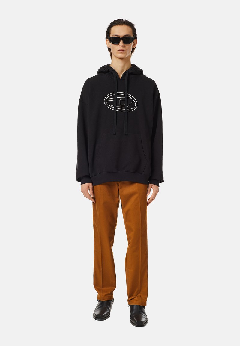 Hoodie noir surdimensionné avec logo blanc, associé à un pantalon droit marron et des bottes noires. Le modèle porte des lunettes de soleil noires. Style minimaliste.