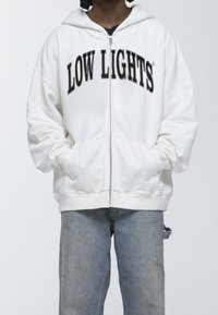 Sudadera blanca con cremallera que presenta "LOW LIGHTS" en texto negro en negrita en el pecho, con capucha y bolsillos frontales. Combinada con jeans de color azul claro.