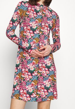 Kvinde iført en langærmet knælængde kjole med et tæt, farverigt blomsterprint i pink, orange, blå og grønne nuancer.