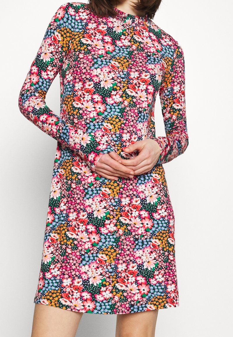 Femme portant une robe à manches longues, longueur genou, avec un motif floral dense et coloré aux tons rose, orange, bleu et vert.