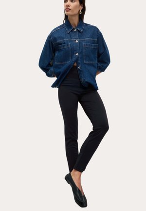 Donna che indossa una giacca di jeans blu scuro, pantaloni neri aderenti, scarpe slip-on nere, in piedi con una gamba incrociata sull'altra.