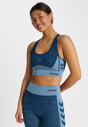 Glimlachende vrouw die een naadloze sport-bh en legging in blauw en lichtblauw draagt, met "hummel"-logo's en chevronpatronen op een witte achtergrond.