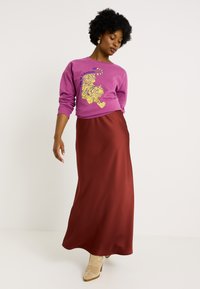 Mujer vestida con sudadera morada que tiene un gráfico de tigre amarillo y texto, combinada con una larga falda roja y botas de tacón beige, de pie contra un fondo blanco.