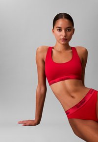 Roter Sport-BH und passende Slips mit schwarzem "Calvin Klein"-Branding auf den elastischen Bändern, glatte Textur und eng anliegende Passform vor einem grauen Hintergrund.
