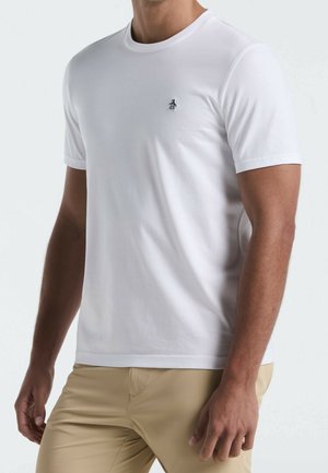 Uomo che indossa una semplice maglietta bianca a maniche corte con un piccolo logo nero di un pinguino sul petto e pantaloni beige.