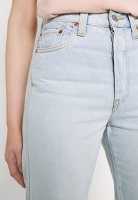 Lähikuva henkilöstä, joka on pukeutunut vaaleansinisiin korkeavyötäröisiin farkkuihin, joissa on kupariset niitit, ja vaaleanpunaiseen toppiin, joka on sisätaskussa.