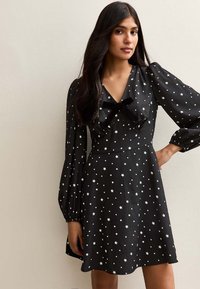 Robe noire aux manches longues bouffantes, avec un col en V, un nœud décoratif et un motif d'étoiles blanches sur tout le tissu.