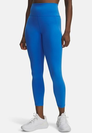 Blaue Leggings mit hohem Bund, aus dehnbarem Material. Sie haben eine glatte Textur, sind bodenlang und besitzen einen schmalen Knöchelofen. Kombiniert mit weißen Turnschuhen.