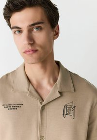 Beige short-sleeve collared shirt featuring black text "LOS ANGELES COUNTY SANTA MONICA SOUNDS" and a graphic of a record.