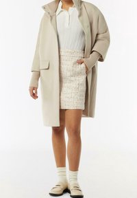 Beige wollen blend jas over een witte geribbelde top en een gestructureerde tweed mini-rok met koraalaccenten, gecombineerd met crème sokken en instap schoenen.