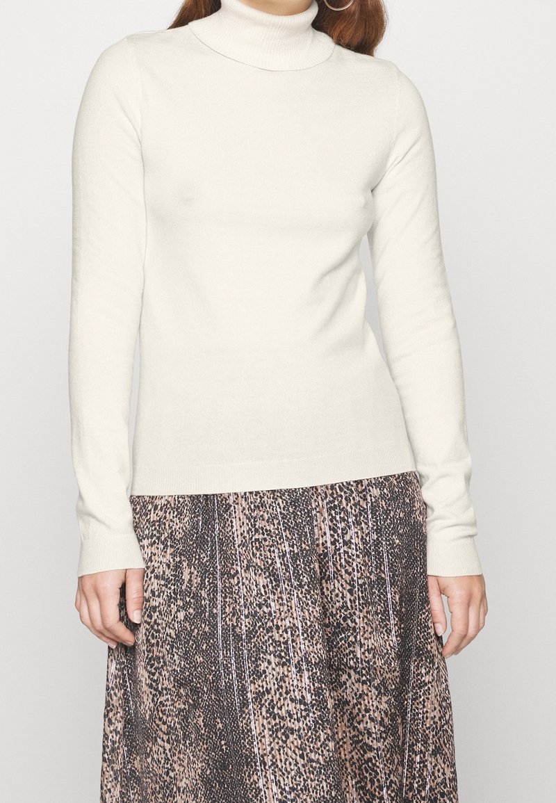 Cream turtleneck sweater in soft fabric, fitted silhouette, long sleeves, with a ribbed texture. Paired with a patterned skirt.