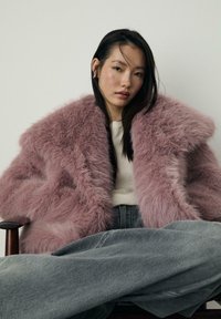 Plašč iz umetnega krzna v mehki roza barvi z velikim ovratnikom, ki se ponaša z puhasto teksturo. Nosite ga preko lahkega puloverja in ohlapnih sivih kavbojk.