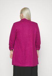 Fuchsia blazer met een gestructureerde afwerking, voorzien van een kraag en gerimpelde mouwen, die voorbij de heupen valt, gecombineerd met een zwarte onderkant.
