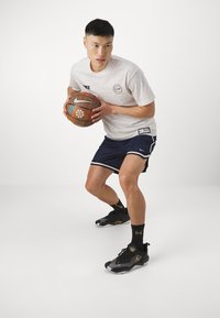 Conjunto atlético de Nike que presenta una camiseta de color claro con patrón y pantalones cortos azul marino con ribetes blancos. Zapatillas de baloncesto negras y doradas, sosteniendo un baloncesto.