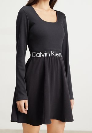 Kvinde iført en sort kjole med lange ærmer med et elastisk bånd med Calvin Klein-logo og en udvidet nederdel mod en ensfarvet baggrund.