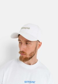 Mann mit rotem Bart, der eine weiße Mütze und einen weißen Sweatshirt mit dem blauen Logo "SIXTHJUNE" trägt, schaut zur Seite vor einem schlichten Hintergrund.