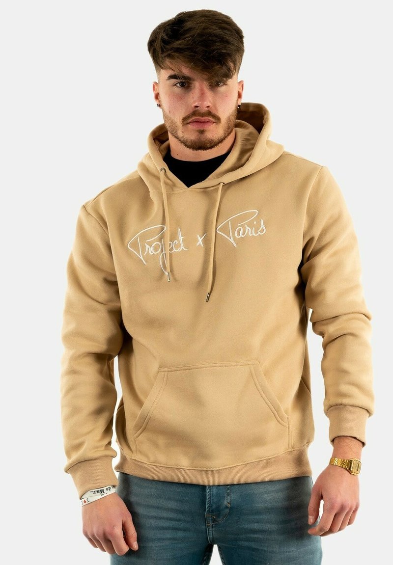 Sudadera beige con capucha y bordado en blanco de "Project x Paris", con bolsillo tipo canguro y puños acanalados. Combinada con jeans azules.
