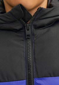 Chaqueta acolchada negra y morada con un alto cuello, que presenta un cierre frontal y costuras azules en contraste a lo largo de las costuras.