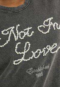 Kogļa pelēkas krāsas T-kreklus ar baltām pērļu pērlītēm veidotu tekstu "Not In Love" un izšūtu detaļu, kas lasa "Established 1985." Mīksta, ikdienišķa auduma.