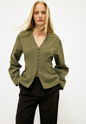 Frau mit langen blonden Haaren trägt eine olivgrüne, geknöpfte Bluse und schwarze Hose, steht mit den Händen in den Taschen vor einfachem Hintergrund.