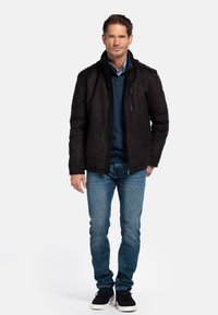 Zwarte suède jacket met een ritssluiting aan de voorkant, staande kraag en zijzakken, gecombineerd met een blauwe trui, jeans en zwarte schoenen.