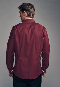 Duga maroon košulja s glatkom teksturom, dugim rukavima, na dugmad na manžetama i ovratniku. Ima zakrivenu donju rub i jedan detalj s dugmetom na vratu.