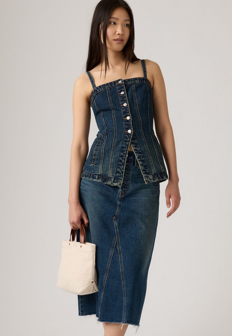 Frau, die ein geknöpftes Oberteil aus dunklem Denim und einen passenden Midirock mit rohem Saum trägt, und eine kleine beigefarbene Tasche mit braunen Griffen hält.