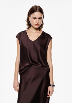 Top in satin marrone con scollatura a V, maniche corte e vestibilità ampia. Il tessuto ha una texture liscia, abbinato a un bracciale nero al polso.