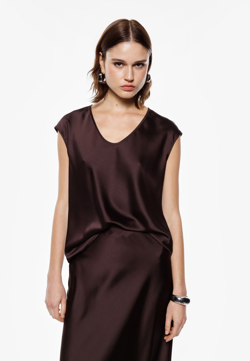 Top in satin marrone con scollatura a V, maniche corte e vestibilità ampia. Il tessuto ha una texture liscia, abbinato a un bracciale nero al polso.