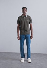 Olīvu zaļš polo krekls ar kontrastainu gaišas krāsas apkakli, kopā ar ziliem džinsiem un baltām sporta apaviem. Viens tonis fons.