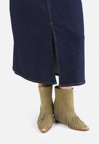 Bottes chelsea en suede vert olive avec des franges et des pointes dorées métalliques, portées avec une jupe en denim bleu marine dotée d'une fente sur le côté.