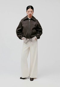 Giacca bomber marrone con colletto alto, chiusura lampo e polsini elasticizzati, abbinata a una camicia bianca oversize e pantaloni a gamba larga.
