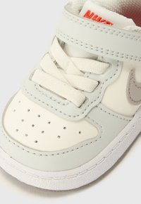 Baskets en cuir gris clair avec des accents beiges, lacets blancs, boîte à orteils perforée et un petit logo swoosh gris sur le côté.
