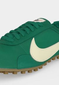 Πράσινο αθλητικό παπούτσι με κρεμ λογότυπο Nike, πράσινα κορδόνια και καφέ υφή σόλας, σε κλίση προς τα δεξιά σε λευκό φόντο.