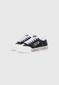 Sorte og hvite joggesko med teksturert overdel, hvit gummisåle og trykt "VERSACE JEANS COUTURE"-logo på siden og hælen.