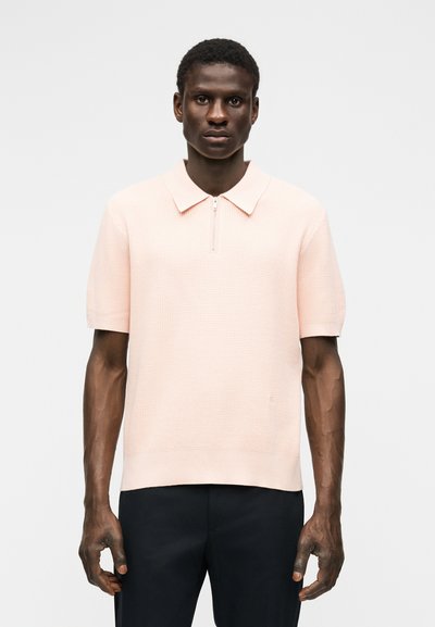 Άνδρας που φοράει ελαφρώς ροζ πλεκτό πόλο με κοντό μανίκι και φερμουάρ στο μπροστινό μέρος, συνδυασμένο με μαύρο παντελόνι, στέκεται μπροστά σε λευκό φόντο.