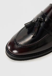 Mocasines de cuero en burdeos con un acabado brillante. Presentan borlas decorativas y costuras en contraste sobre una suela texturizada negra.