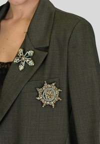 Blazer verde scuro con due spille: una stella a raggio in argento e oro con pietre chiare e una spilla geometrica con accenti dorati e dettagli verdi.