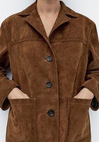 Brązowa zamszowa kurtka z lekkim połyskiem, z klapowym kołnierzem, dwoma dużymi przednimi kieszeniami i trzema czarnymi guzikami z przodu.